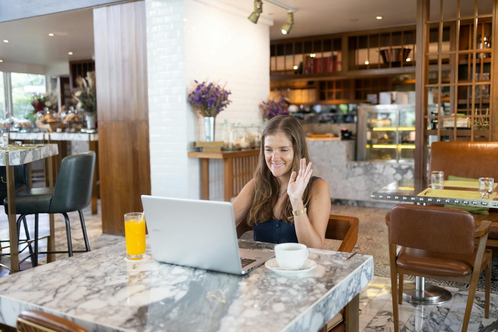 Woman smiling and waving during video call at a chic cafe with a marble table.